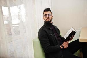 un homme arabe porte une veste en jean noir et des lunettes assis dans un café, lit un livre. mec modèle arabe élégant et à la mode. photo