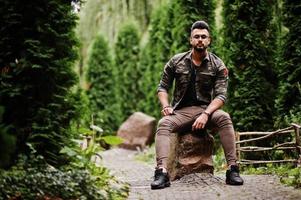 Superbe homme macho à barbe arabe de grande taille dans des lunettes et une veste militaire posée à l'extérieur, assis sur la pierre. photo