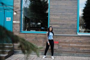 fille afro-américaine hipster portant une chemise en jean à manches léopard posant dans la rue contre une maison en bois avec des fenêtres. photo