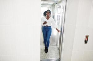 femmes afro-américaines élégantes en chemisier blanc et jeans bleus posés à l'ascenseur avec un téléphone portable à portée de main. photo