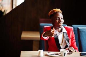 modèle d'homme afro-américain de mode en costume rouge, avec des cheveux en surbrillance assis au café avec du café et vous montrer le doigt. photo