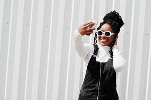 femme africaine en jupe salopette en jean, lunettes de soleil blanches posées contre un mur en acier blanc et faisant du selfie par téléphone. photo