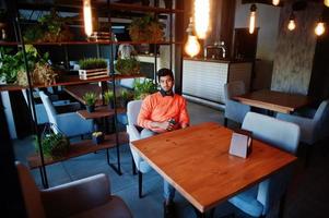 jeune homme indien confiant dans des vêtements décontractés intelligents comme un pull orange, assis au café et utilisant son téléphone portable. photo