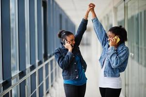 deux amies africaines en veste de jeans parlent au téléphone et mains en l'air à l'intérieur ensemble. photo