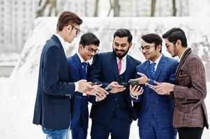 groupe de cinq hommes d'affaires indiens en costumes posés en plein air en journée d'hiver en europe, regardant sur les téléphones et disscus. photo