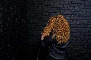 fille aux cheveux bouclés à la veste en cuir sur studio contre le mur de briques noires. photo