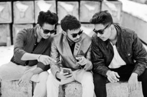 groupe de trois jeunes indiens décontractés portant des lunettes de soleil posées contre des blocs de pierre et regardant un téléphone portable. photo