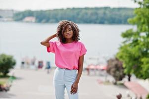 une fille bouclée afro-américaine posée dans la rue de la ville porte un chemisier rose et un pantalon bleu. photo