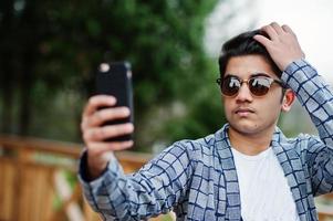 élégant jeune homme indien à lunettes de soleil porter décontracté posé en plein air et faire selfie au téléphone mobile. photo