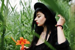 fille sensuelle tout en noir, lèvres rouges et chapeau. une femme dramatique gothique tient une fleur de lys orange sur un roseau commun. photo