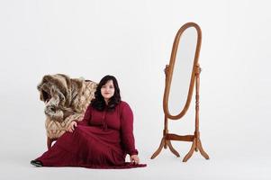 jolie femme sud-asiatique en robe rouge foncé posée au studio sur fond blanc contre le miroir et la chaise. photo