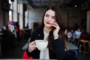 fille brune assise sur un café avec une tasse de cappuccino et parlant de téléphone portable. photo