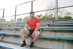 jeune homme musclé barbu brutal portant une chemise rouge, un short et une casquette au stade. photo