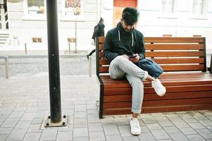 portrait d'un jeune mannequin indien élégant pose dans la rue, assis sur un banc et tient le smartphone à portée de main. photo