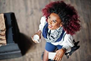 jolie fille bouclée afro-américaine en blouse blanche et short bleu posé au café avec latte et téléphone portable à portée de main. vue d'en-haut. photo