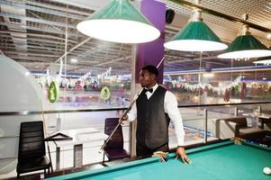 jeune bel homme africain vêtu d'une chemise blanche, d'un gilet noir et d'un nœud papillon jouant au billard. photo