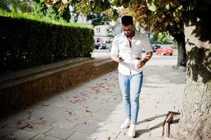 grand modèle d'homme arabe élégant en chemise blanche, jeans et lunettes de soleil marchant dans la rue de la ville. barbe arabe attrayant avec une tasse de café et un téléphone portable à portée de main. photo