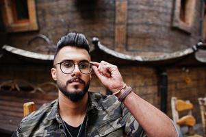 portrait en gros plan d'un magnifique et grand homme macho à la barbe arabe dans des lunettes et une veste militaire posée à l'extérieur, assis sur une chaise contre une maison en bois. photo