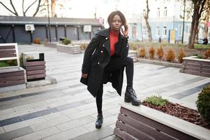 portrait d'une femme africaine aux cheveux bouclés portant un manteau noir à la mode et un col roulé rouge posant à l'extérieur. photo