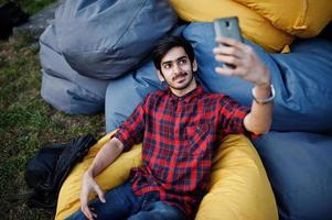 jeune étudiant indien en chemise à carreaux et jeans assis et se détendre sur des oreillers extérieurs et faire du selfie. photo