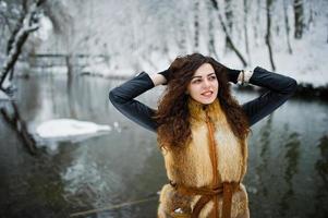 élégance fille bouclée en manteau de fourrure au parc forestier enneigé contre la rivière gelée en hiver. photo