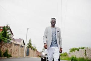 homme afro-américain riche et élégant en blazer et pantalon blanc, lunettes posées contre une voiture suv. photo