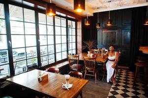 femme afro-américaine, coiffure rétro en robe blanche au restaurant avec verre de vin. photo