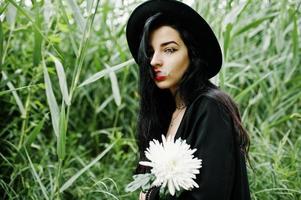 sensuelle fille fumeuse tout en noir, lèvres rouges et chapeau. une femme dramatique gothique tient une fleur de chrysanthème blanche et fume sur un roseau commun. photo