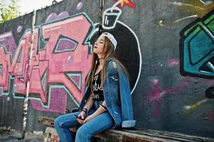 une fille hipster décontractée et élégante en casquette et en jean porte de la musique à partir d'un casque de téléphone portable contre un grand mur de graffitis avec une bombe. photo