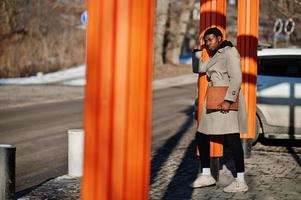 bel homme afro-américain posant à l'extérieur avec un chapeau noir et un manteau beige avec un dossier à la main. photo