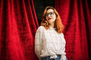 jolie femme rousse à lunettes, porter sur un chemisier blanc posant à l'arche de rideaux rouges ouverts. photo