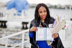 étudiante afro-américaine en robe de graduation noire avec diplôme, au costume bleu posé. photo