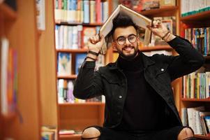 grand étudiant arabe intelligent, portant une veste en jean noir et des lunettes, assis à la bibliothèque et se couvrant la tête avec un livre. photo