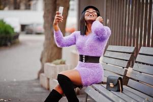 femme afro-américaine en robe violette et casquette posée à l'extérieur, assise sur un banc et faisant un selfie au téléphone. photo