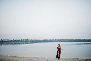 femme aux pieds nus sensuelle blonde en robe de marsala rouge posant contre le lac sur le sable. photo