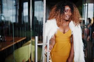 femme afro-américaine glamour en robe jaune et cape en laine blanche avec sac à main posé à l'entrée de la porte vitrée du restaurant. photo