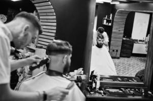 jeune homme barbu se coupe les cheveux par un coiffeur assis sur une chaise au salon de coiffure. âme de barbier. photo