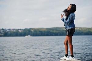 modèle mince afro-américain à la peau foncée posé dans un short noir et une veste en jean contre le bord de mer. photo