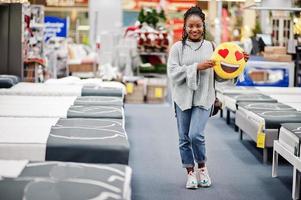 face à l'emoji coeur d'amour. femme africaine avec oreiller dans un magasin d'ameublement moderne. photo