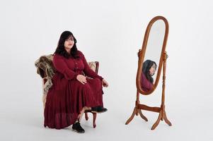 jolie femme sud-asiatique en robe rouge foncé posée au studio sur fond blanc contre le miroir et assis à la chaise. photo