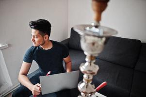 homme indien beau et à la mode en noir assis dans la chambre, fumant du narguilé et travaillant sur un ordinateur portable. photo