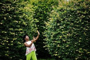 incroyable femme modèle afro-américaine en pantalon vert et chapeau noir posé au parc. photo