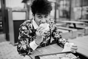 attrayant et beau homme indien élégant porter sur la chemise manger un hamburger et des frites sur la terrasse du café-salon moderne à sa pause de travail. photo