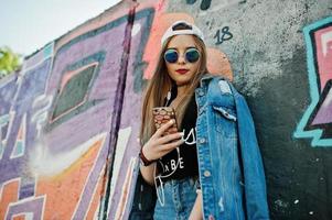 élégante fille hipster décontractée en casquette, lunettes de soleil et jeans, écoutant de la musique à partir d'un casque de téléphone portable contre un grand mur de graffitis. photo