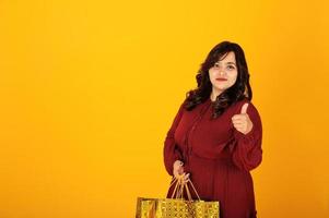 jolie femme sud-asiatique en robe rouge foncé posée au studio sur fond jaune avec des sacs à provisions dorés. photo