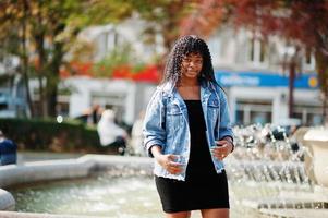 femme bouclée afro france élégante et branchée posée au jour de l'automne en veste de jeans. modèle féminin africain noir contre la fontaine. photo