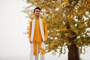 homme élégant indien en vêtements traditionnels jaunes avec foulard blanc posé en plein air contre l'arbre des feuilles d'automne. photo