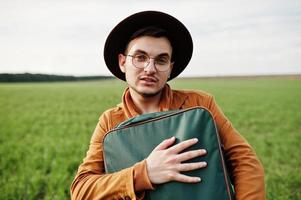 homme élégant à lunettes, veste marron et chapeau avec sac posé sur champ vert. photo