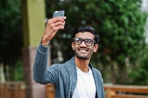 un homme indien élégant à lunettes porte une pose décontractée en plein air et fait du selfie au téléphone. photo