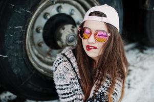 brune élégante fille décontractée en casquette et lunettes de soleil assise contre les roues du camion. photo